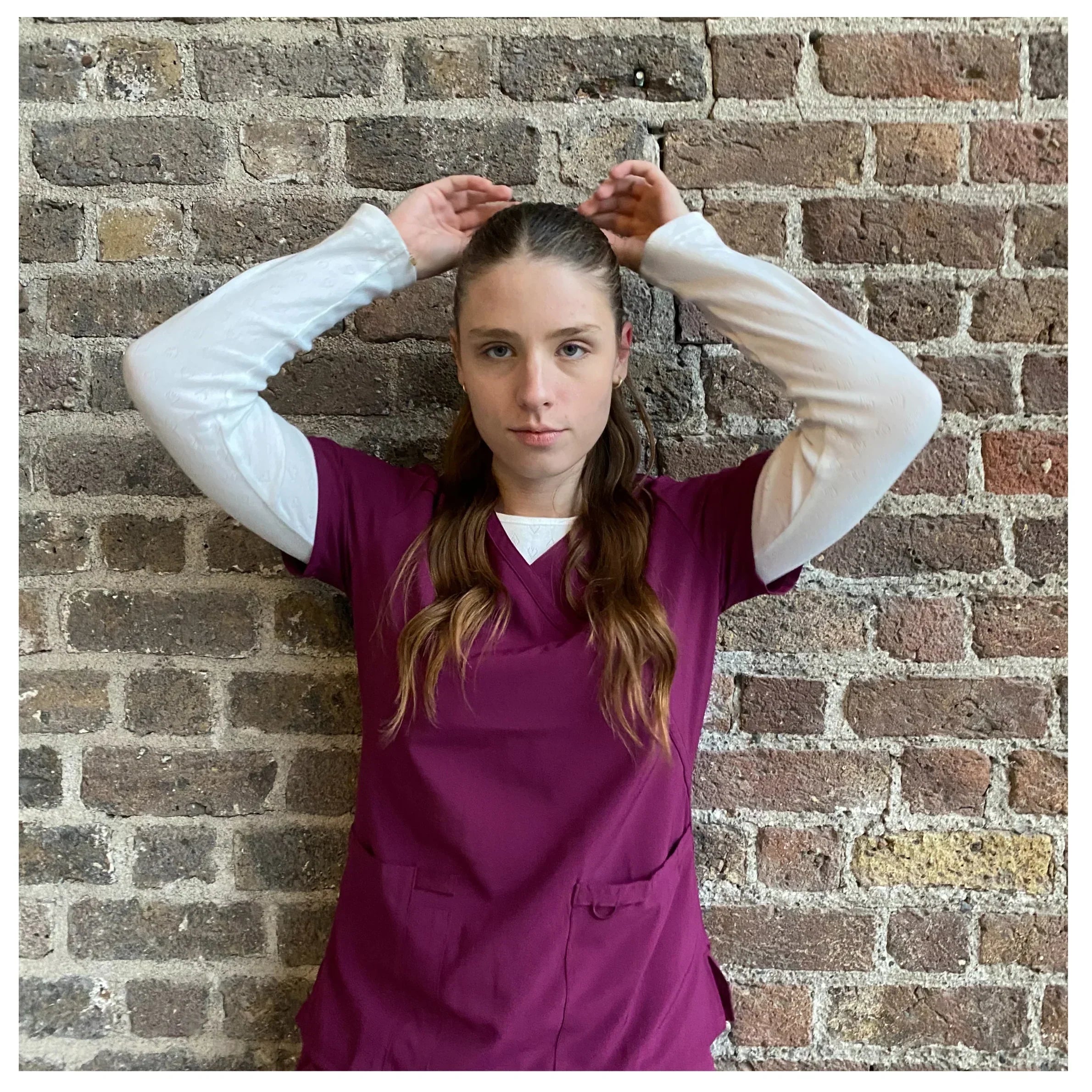 Nurse practitioner wearing burgundy medical scrubs layered over a white long-sleeve top, standing against a brick wall and adjusting her hair, presenting a clean and professional clinical look.