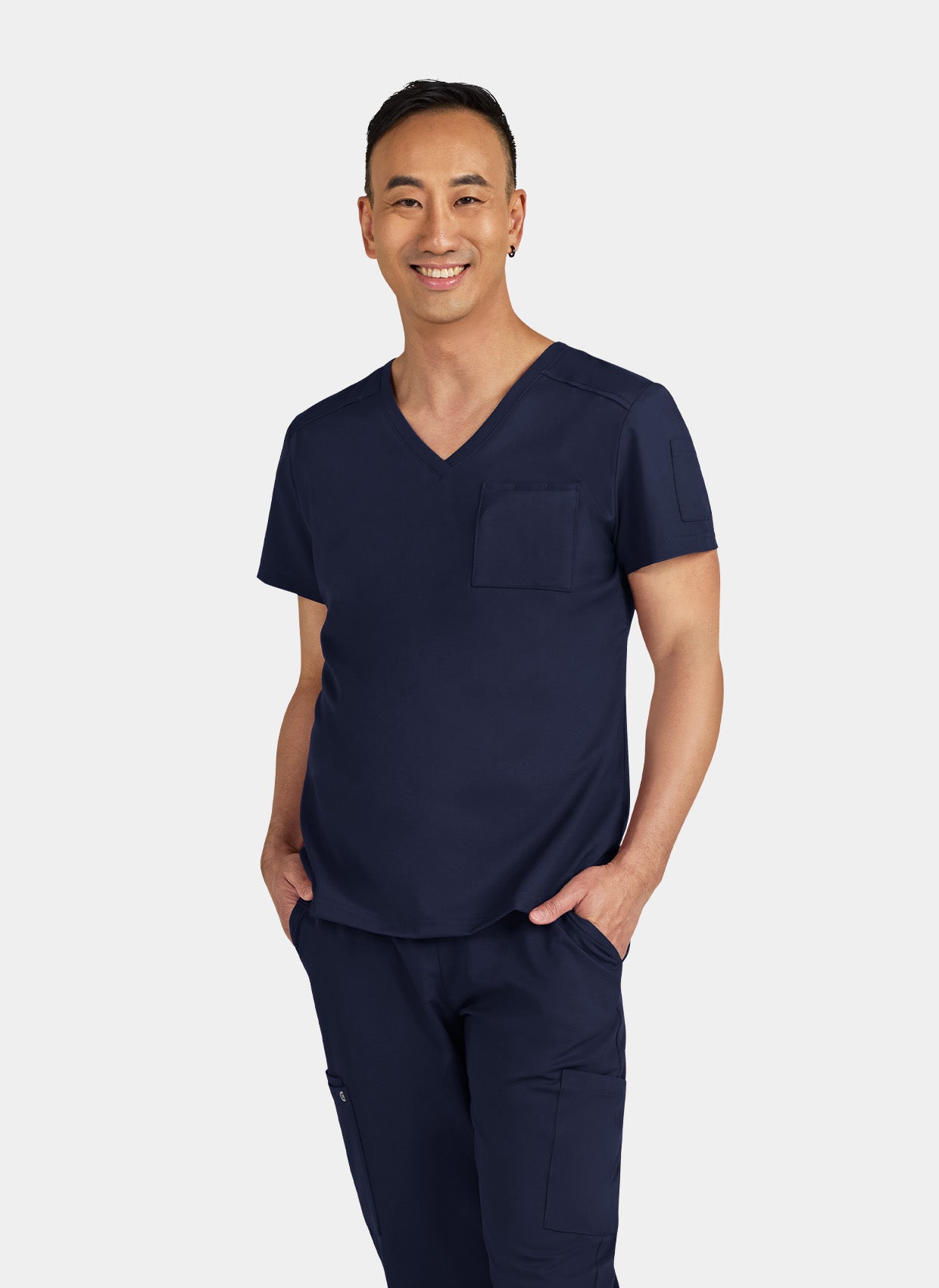 Man wearing navy V-neck scrub top and matching scrub trousers with cargo pockets, hands in pockets.
