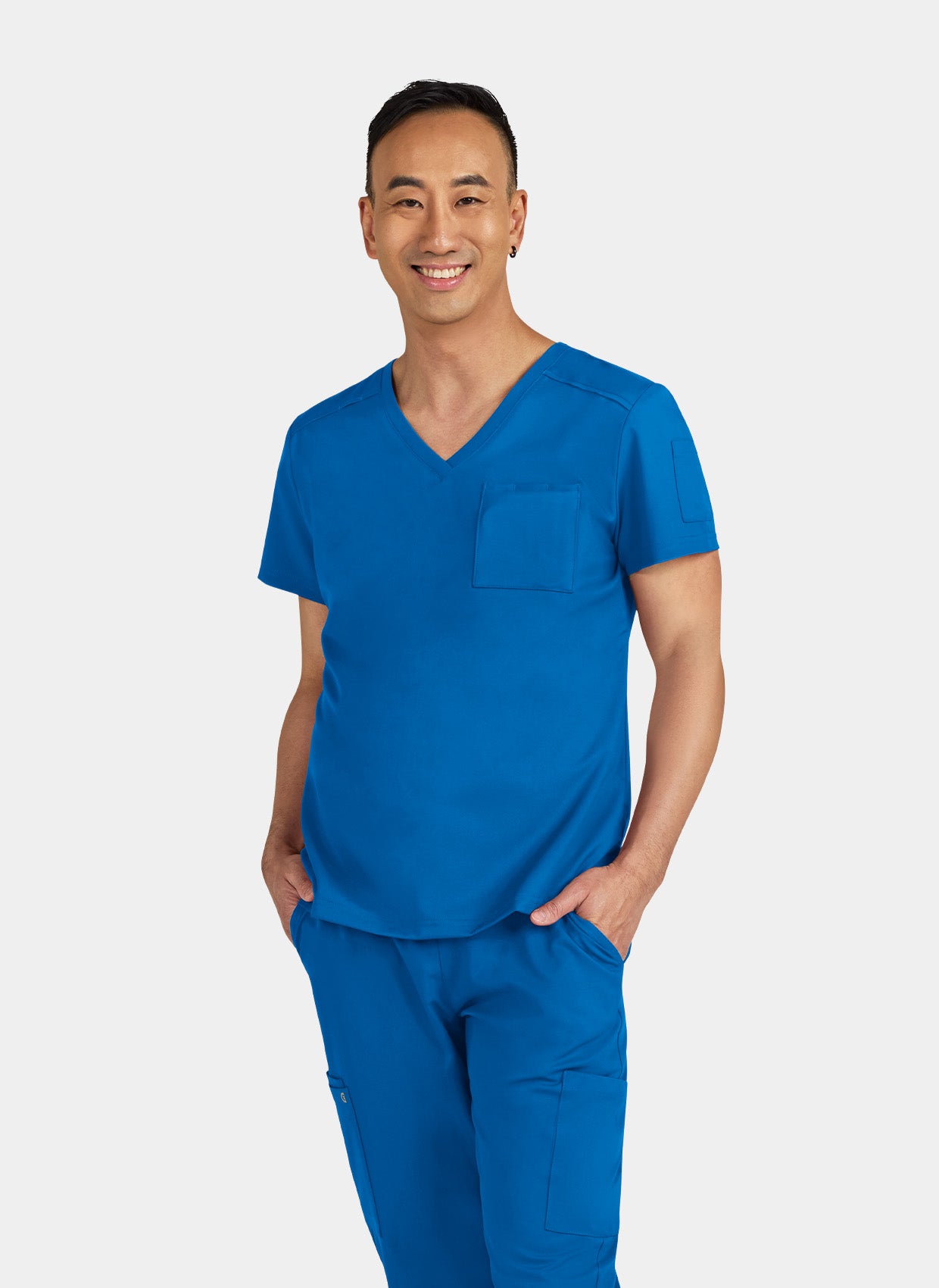 Man wearing a royal blue V-neck scrub top and matching scrub trousers with cargo pockets, hands in pockets.
