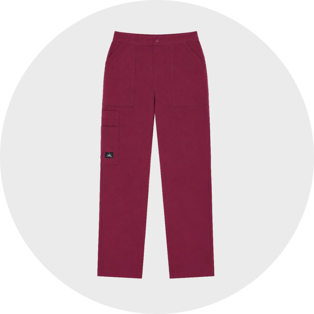 Burgundy straight-leg koi scrub trousers with a button closure, angled front pockets, large thigh pockets, and a small logo patch on the left leg, shown flat against a light gray circular background.